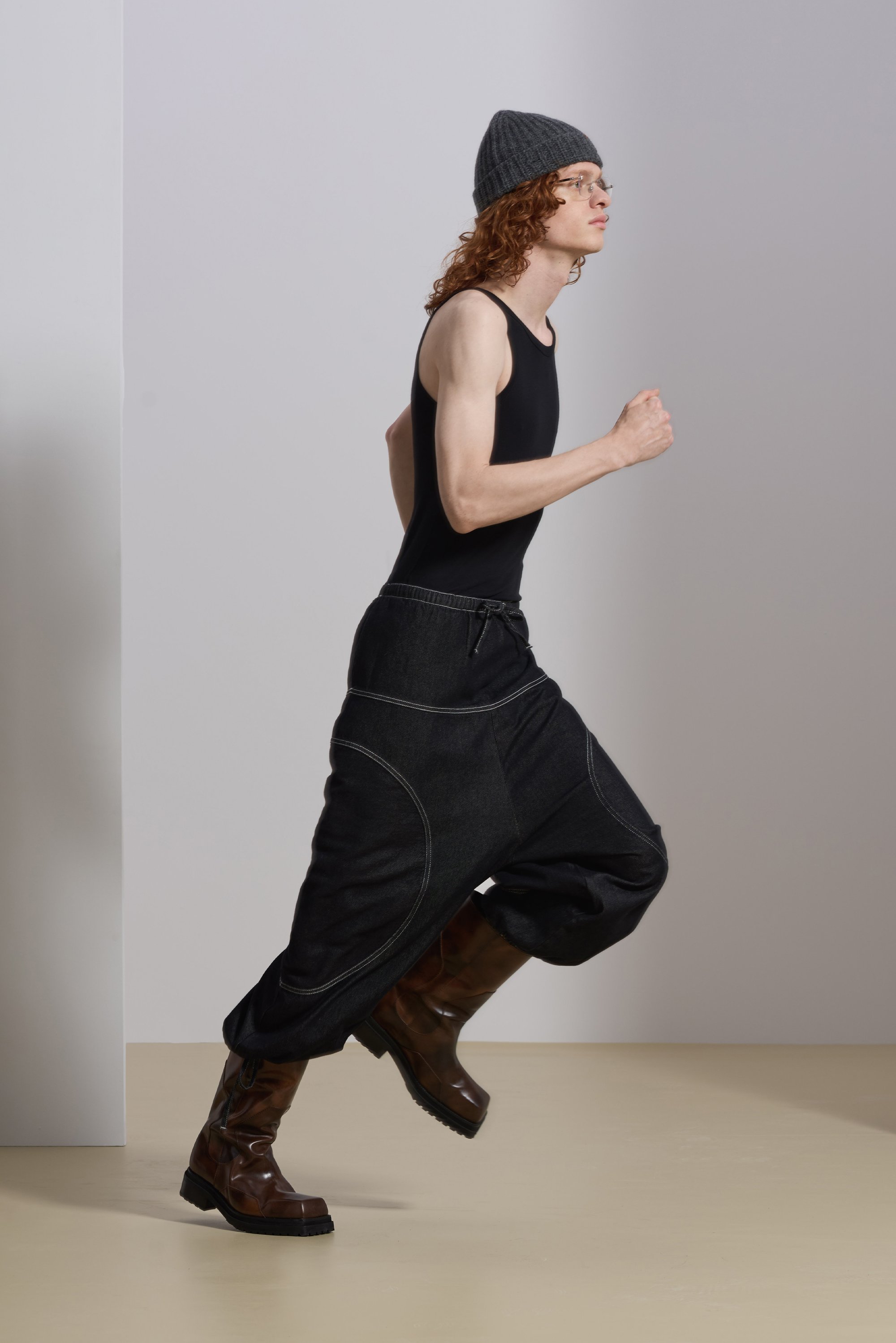 Black denim pants with curved seams, a black tank top, and brown knee-high boots. The figure is wearing a gray knit beanie and glasses.