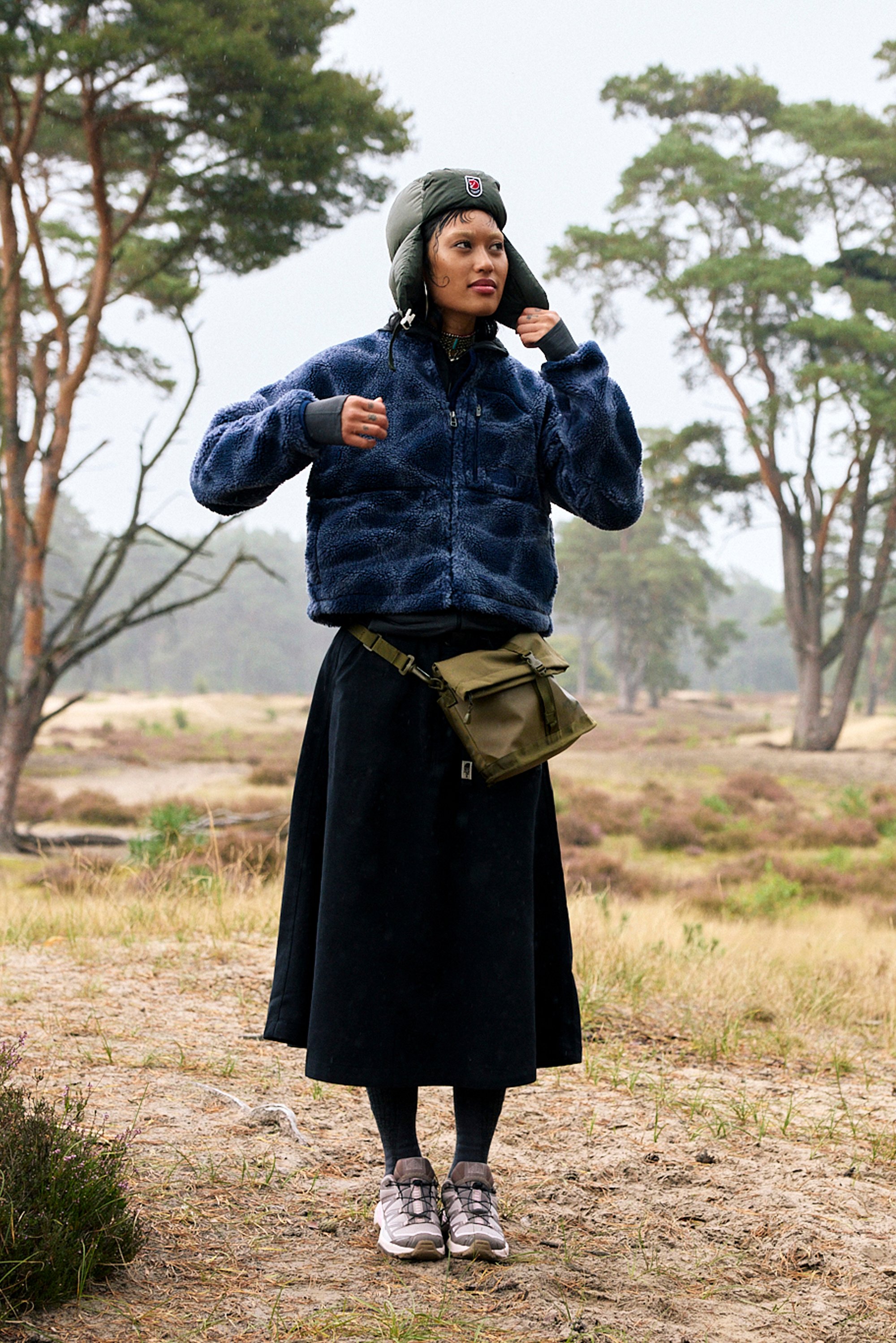 Navy fleece jacket, black skirt, green crossbody bag, grey sneakers, textured materials, outdoor setting with trees in the background.