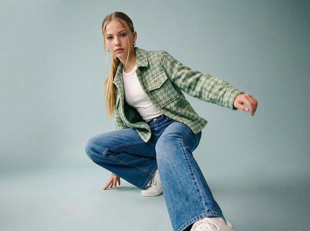 Jeune femme accroupie avec une main posée au sol, portant une veste verte, un haut blanc, un jean bleu et des baskets blanches sur un fond uni.