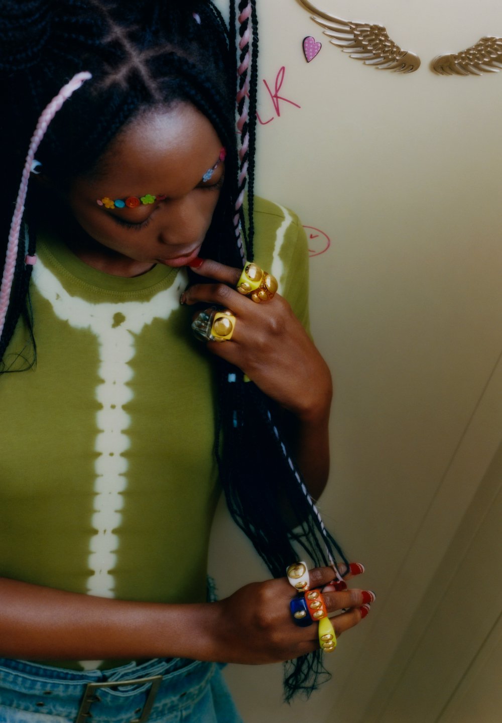 Persona joven con trenzas intrincadas y horquillas de colores lleva una camiseta verde y múltiples anillos llamativos, tocándose suavemente el cabello.