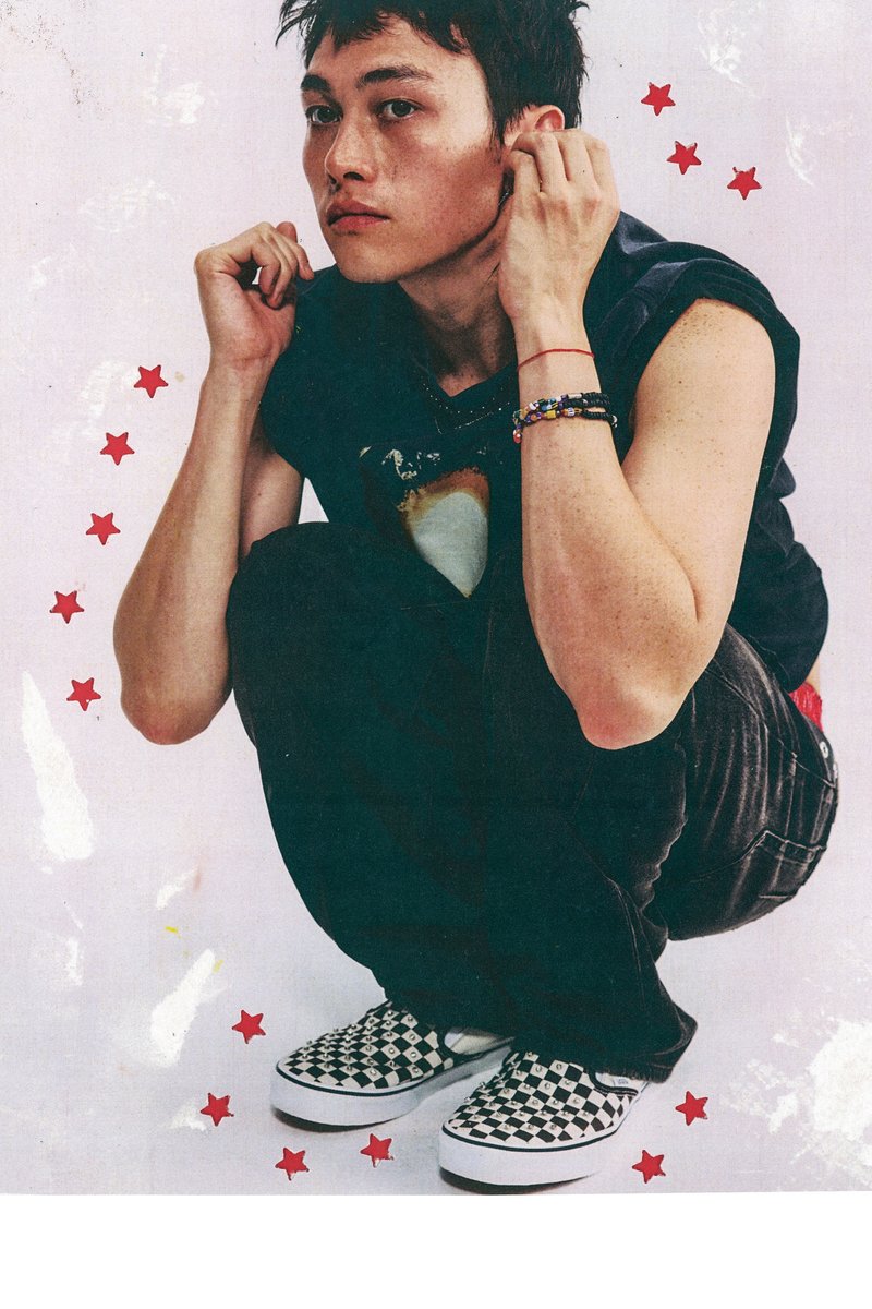Young man crouching, wearing a sleeveless black shirt, checkered slip-on shoes, and colorful bracelets, surrounded by small red star graphics.