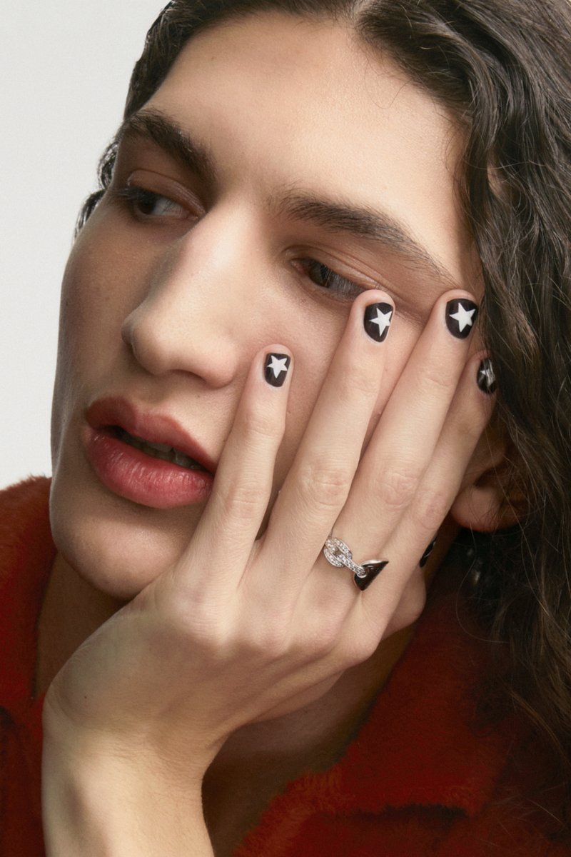Una persona con capelli lunghi e mossi appoggia il mento sulla mano, mettendo in mostra un nail art a motivo stellato e un anello decorativo.