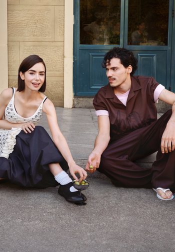 Nuori mies ja nainen istuvat jalkakäytävällä sinisen oven lähellä, jakavat oliiveja pienestä kulhosta, nainen hymyilee, mies katsoo häntä.