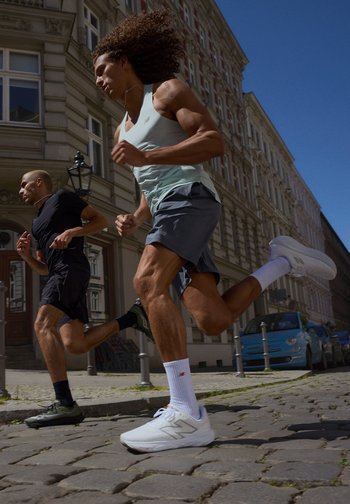 Deux hommes courent rapidement sur une rue pavée, portant des tenues et des chaussures de sport. Un bâtiment historique et des voitures garées se trouvent en arrière-plan.