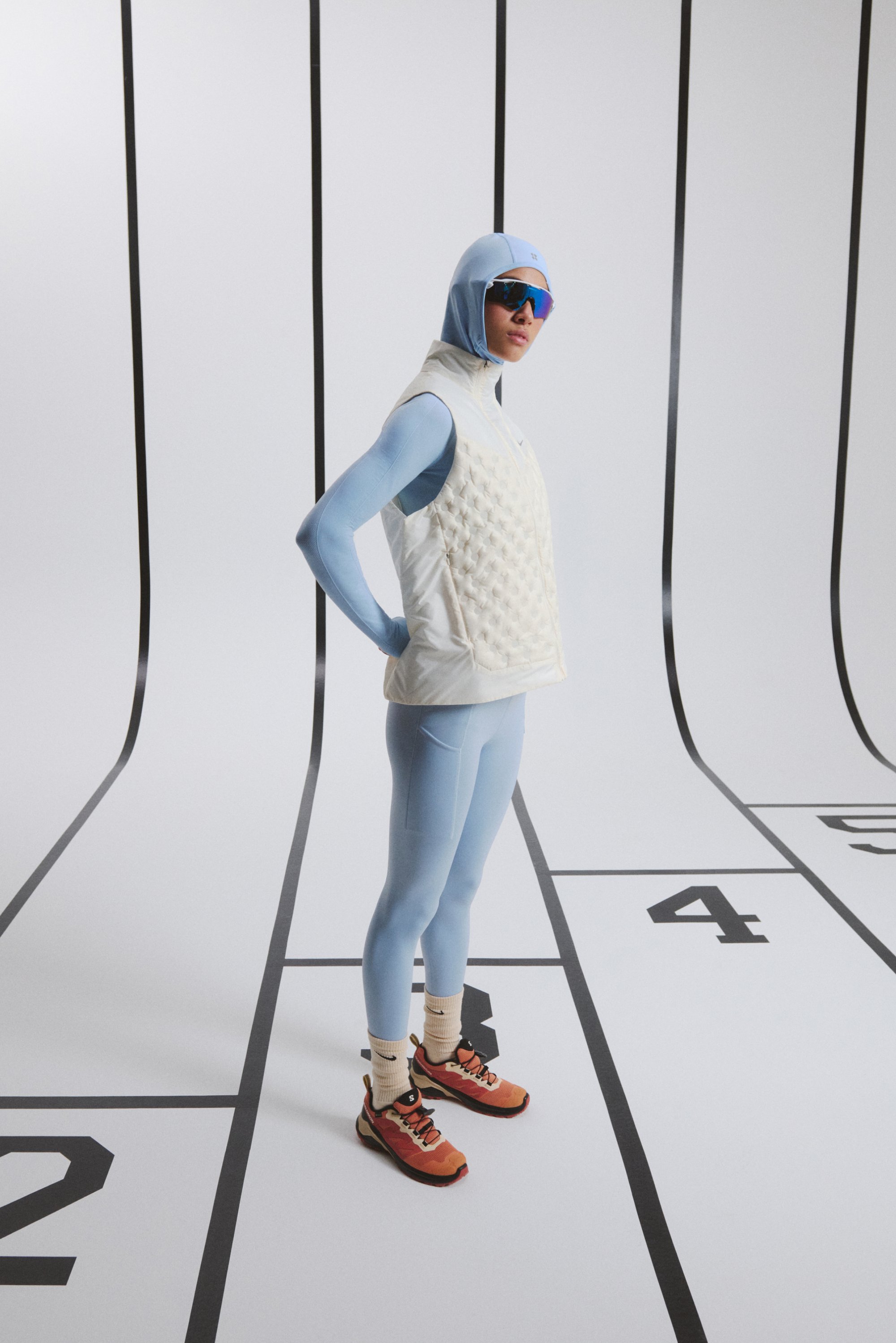 Una persona se encuentra de pie con confianza en un conjunto entallado de color azul claro y un chaleco acolchado, usando gafas de sol en una pista con carriles numerados al fondo.