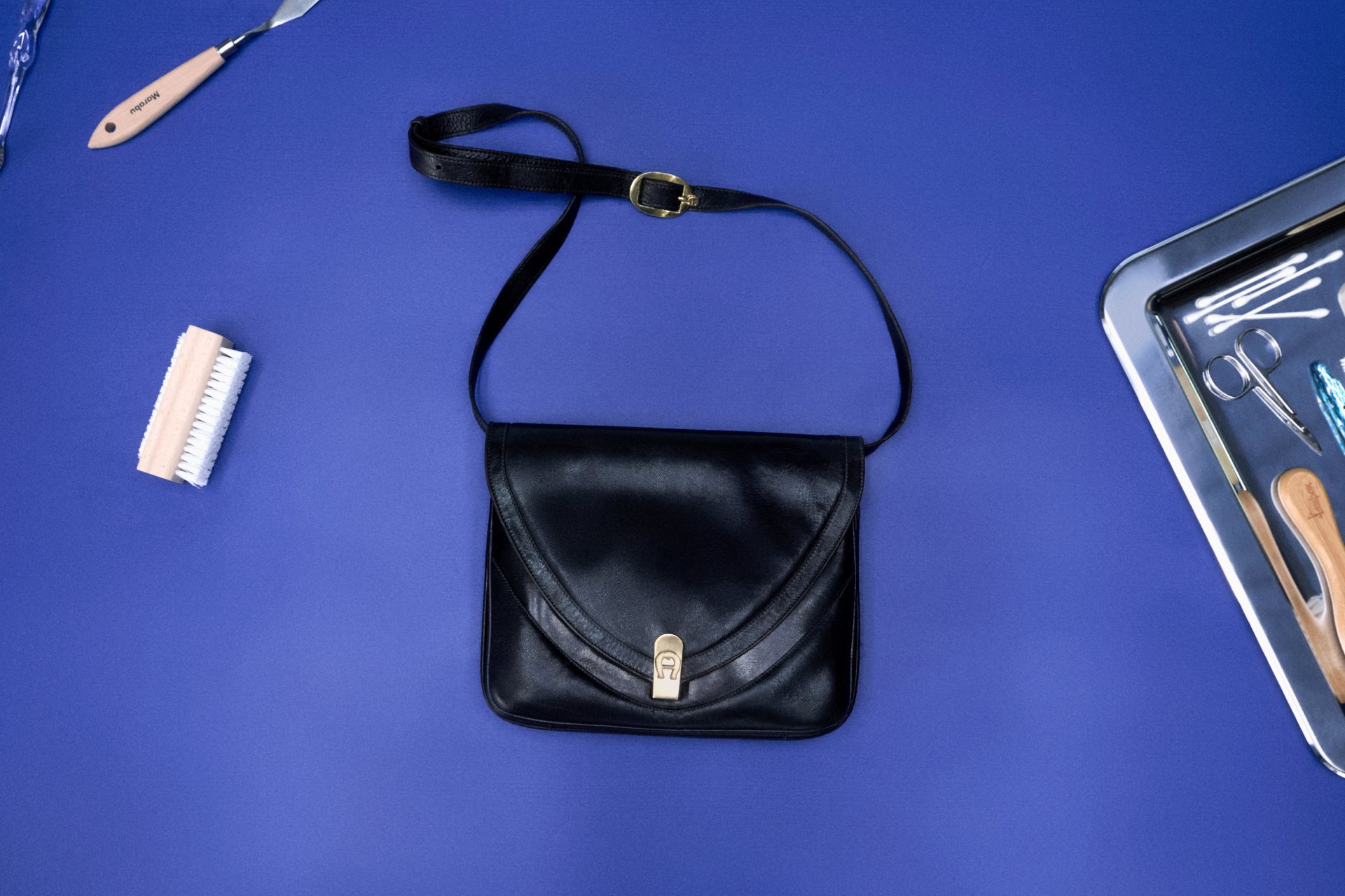 Borsa a tracolla in pelle nera centrata su una superficie viola, accompagnata da una spazzola in legno, attrezzi e un vassoio con vari oggetti.