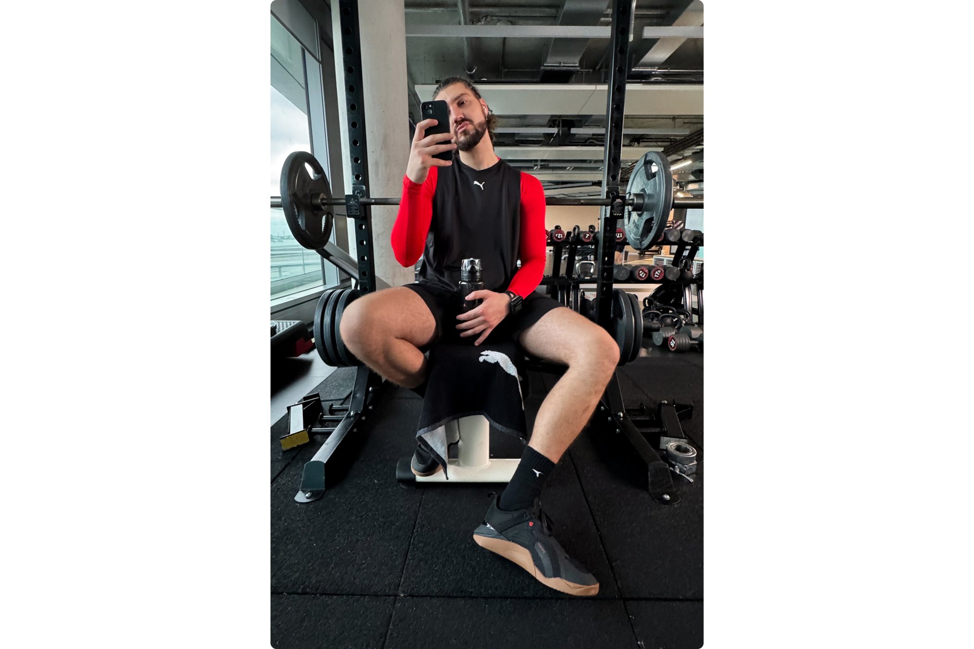 Un hombre en un gimnasio está sentado en un banco de pesas, sosteniendo una botella de agua y un teléfono, vistiendo una camiseta de manga larga roja y pantalones cortos negros. Se pueden ver discos de pesa.