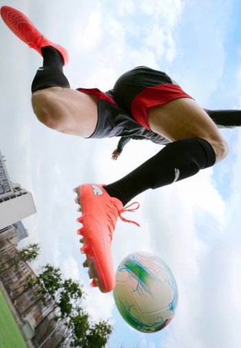 Voetballer in zwart-rode korte broek trapt een wit-blauwe voetbal, draagt feloranje voetbalschoenen en zwarte sokken op een buitenveld.