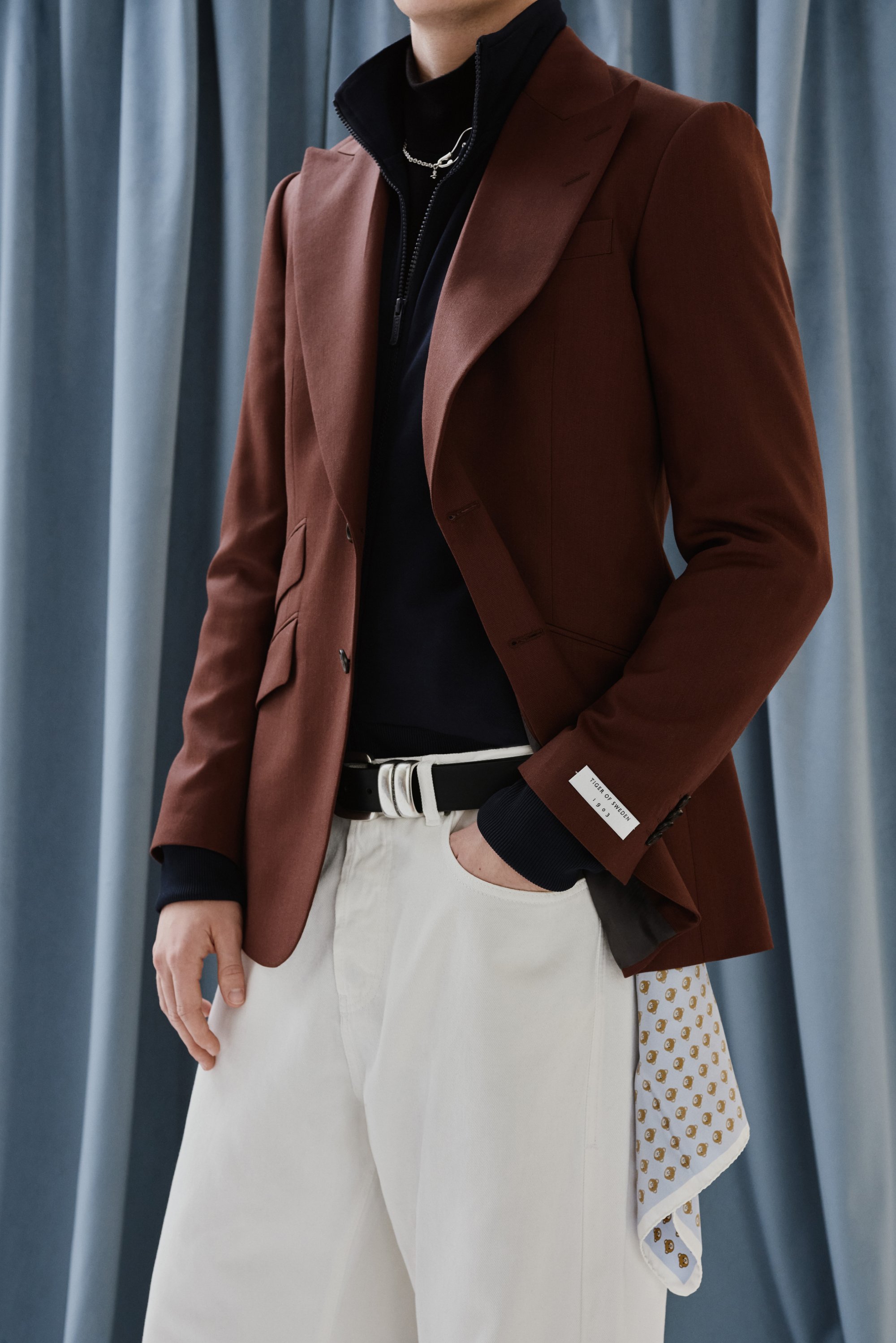 A person models a brown blazer over a black zip-up shirt, paired with white trousers. A patterned pocket square peeks from the blazer.