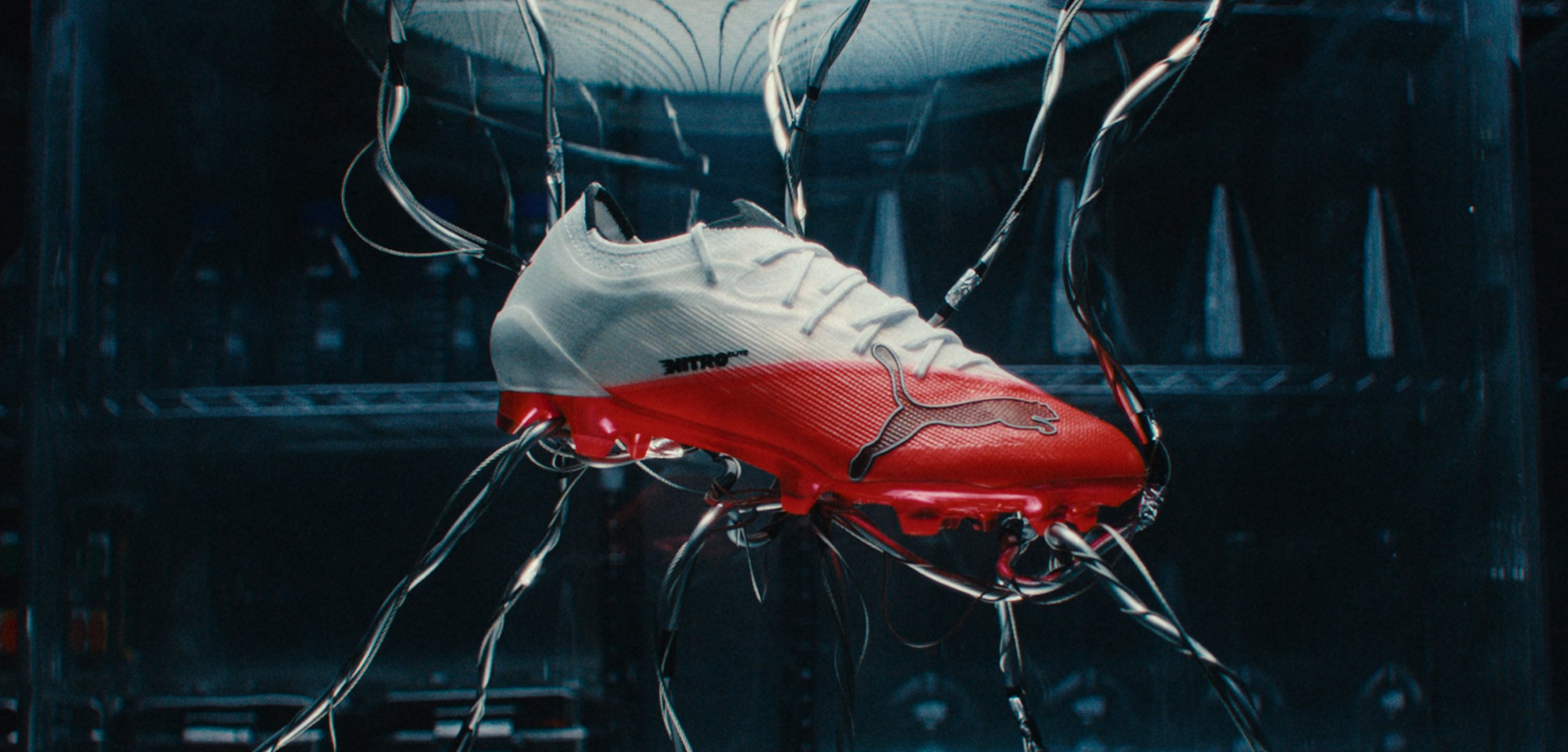 Weiß-roter Puma Fußballschuh, der mit mehreren schwarzen Kabeln in einer dunklen Test- oder Ausstellungsumgebung aufgehängt ist.