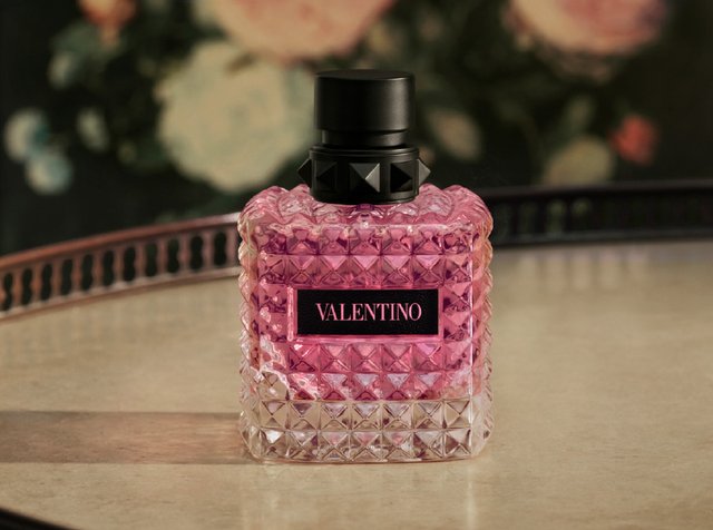 Przezroczysta szklana buteleczka perfum Valentino z różowym płynem i czarną zakrętką na odbijającej powierzchni z kwiatowym tłem.