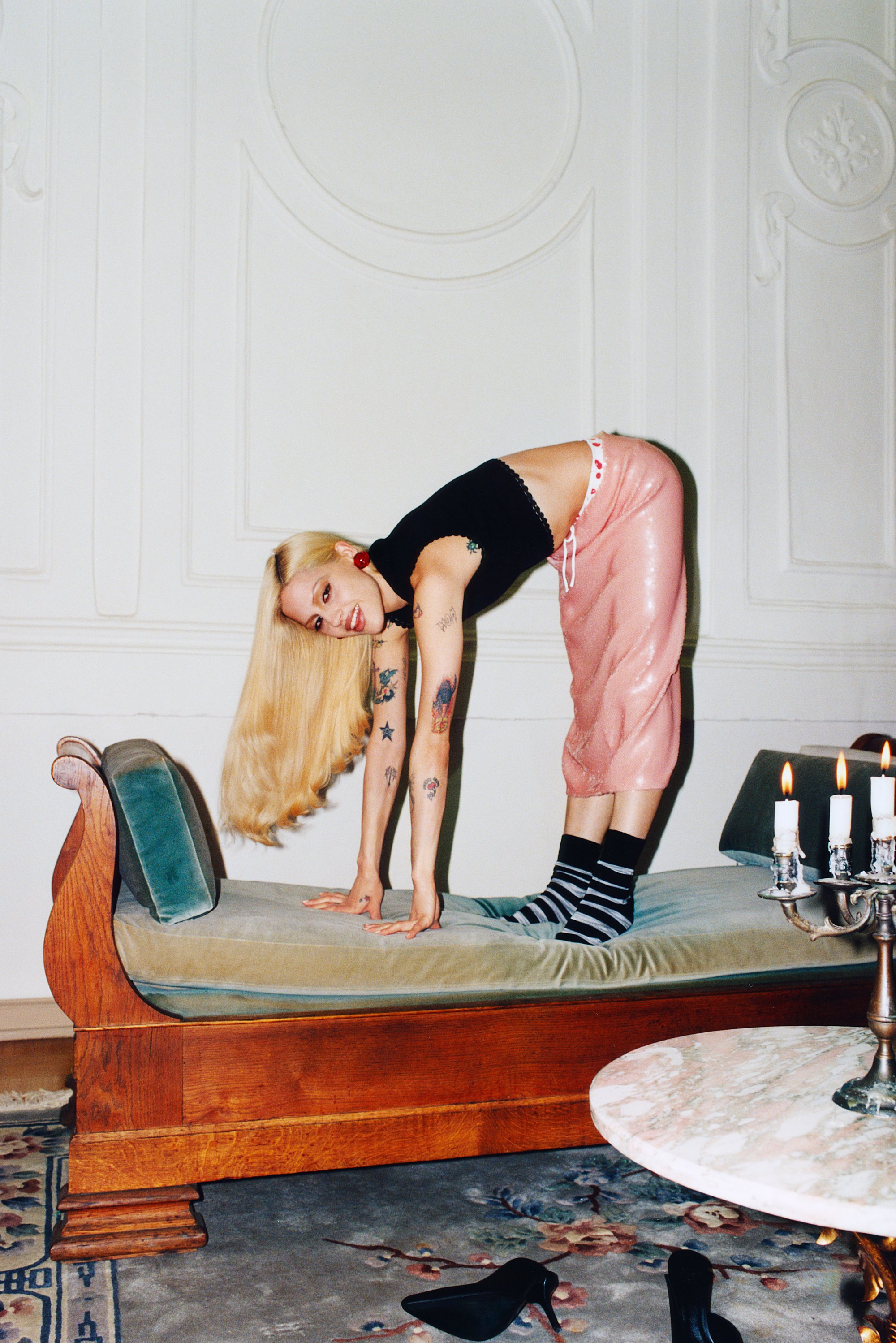 A person with long blonde hair and tattoos performs a playful pose on a couch, wearing pink pants and striped socks, with candles nearby.