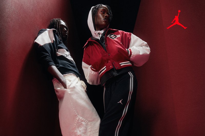 Zwei junge Personen posieren in sportlicher urbaner Kleidung mit dem Jordan-Logo vor roten Wänden und einem schwarzen Hintergrund.