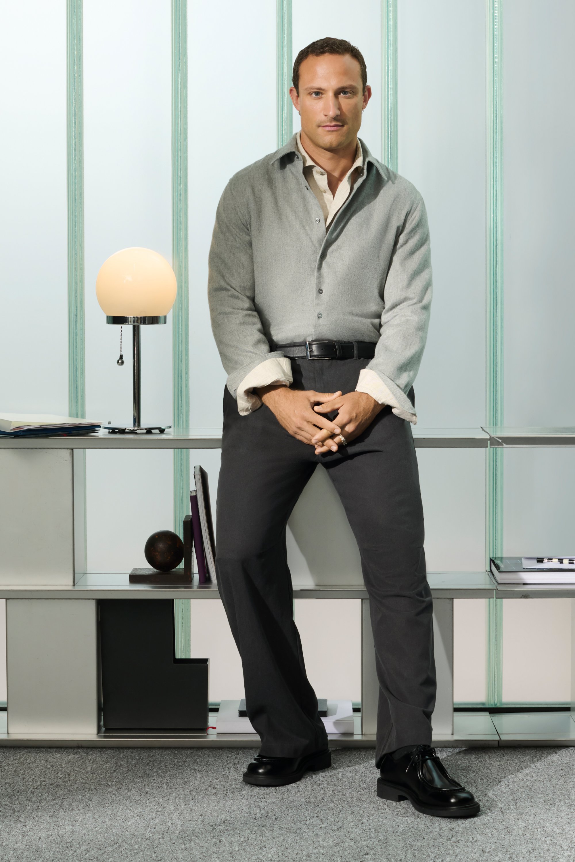 Camicia grigia chiara con polsini bianchi, pantaloni grigio scuro e scarpe nere. In piedi accanto a una lampada da tavolo su un moderno scaffale.