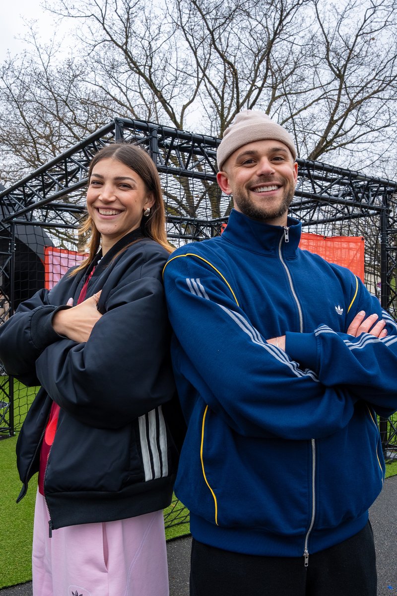 Smiling man and woman stand back-to-back with arms crossed wearing casual jackets outdoors near black metal playground structure.