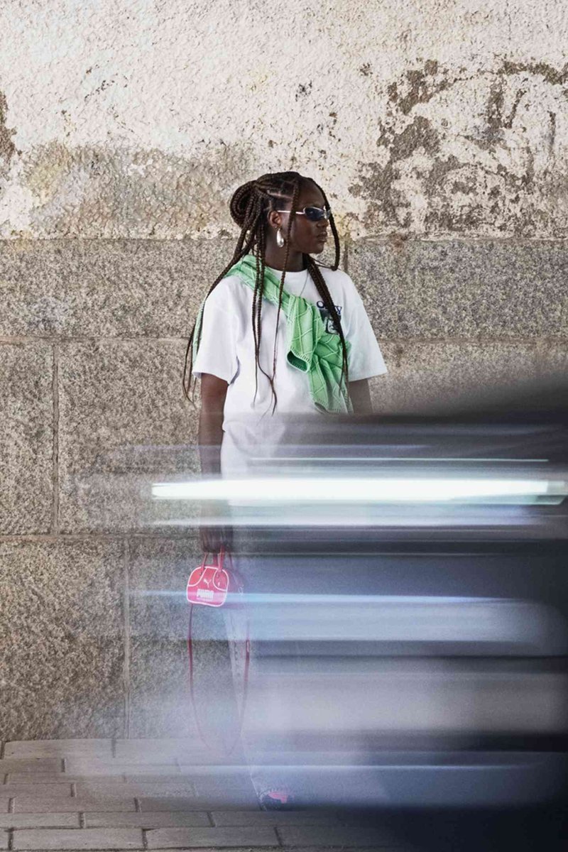T-shirt blanc avec une écharpe à motifs verts, jeans bleus, boucles d'oreilles créoles et sac à main rouge ; debout contre un mur texturé avec une voiture floue en mouvement.