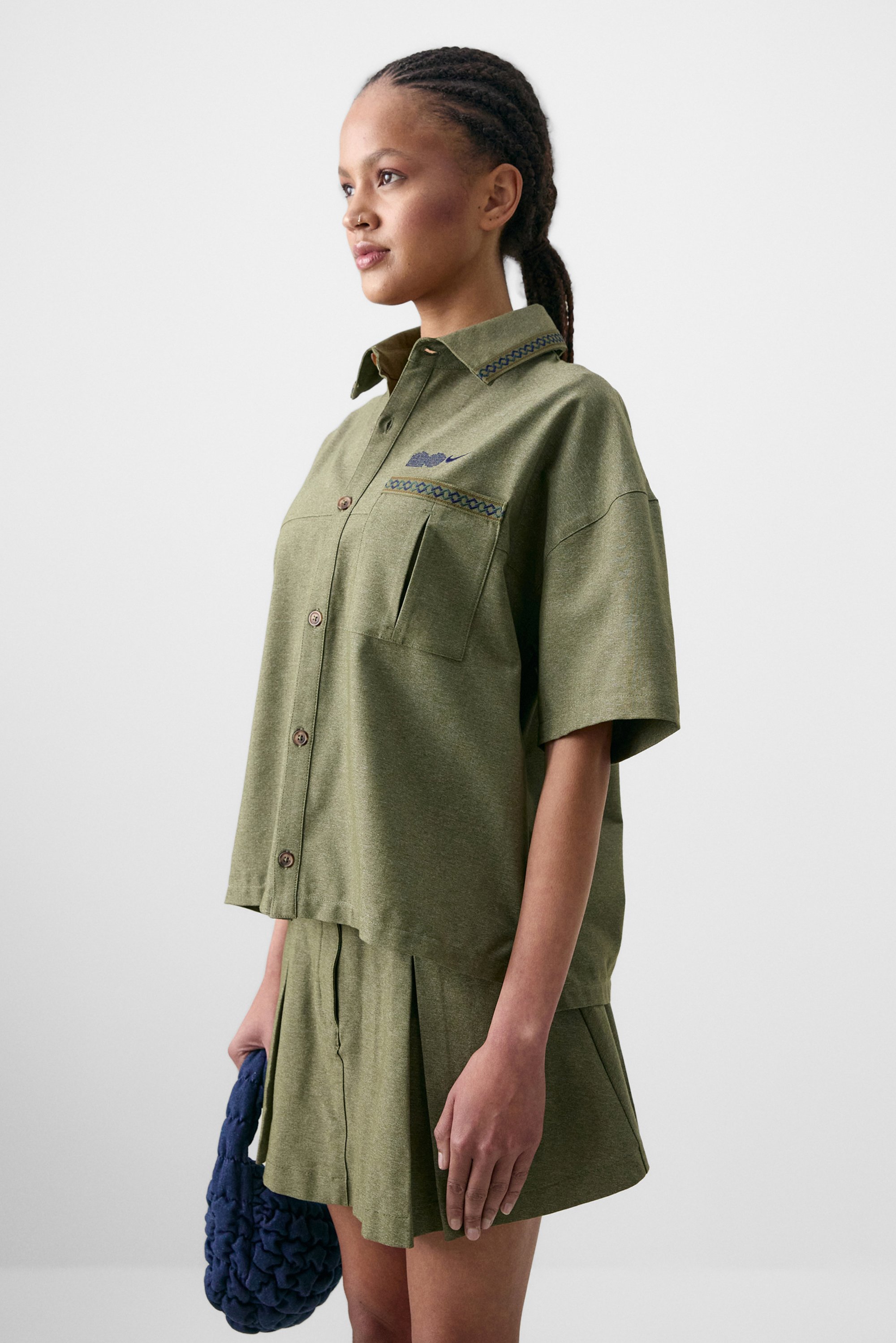 La modelo posa de lado, vistiendo una camisa verde de gran tamaño con un bolsillo y una falda a juego, sosteniendo un bolso de mano tejido azul contra un fondo claro.