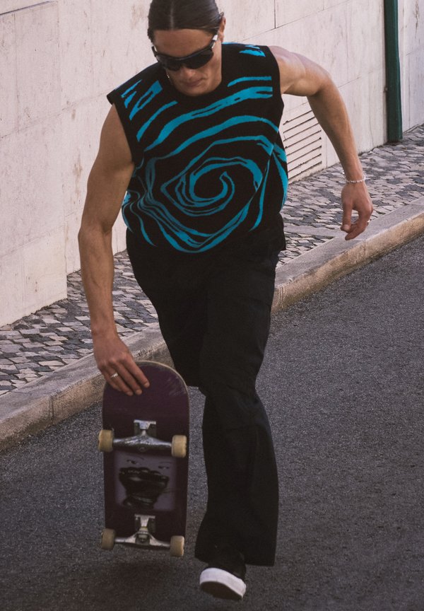 Man wearing sunglasses and a sleeveless black shirt with blue spiral design, holding a skateboard while walking on a paved street.