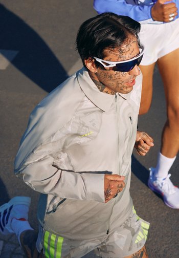 Tatoveret mand med solbriller og let løbetøj jogger udendørs sammen med en anden løber i hvide shorts og sneakers.