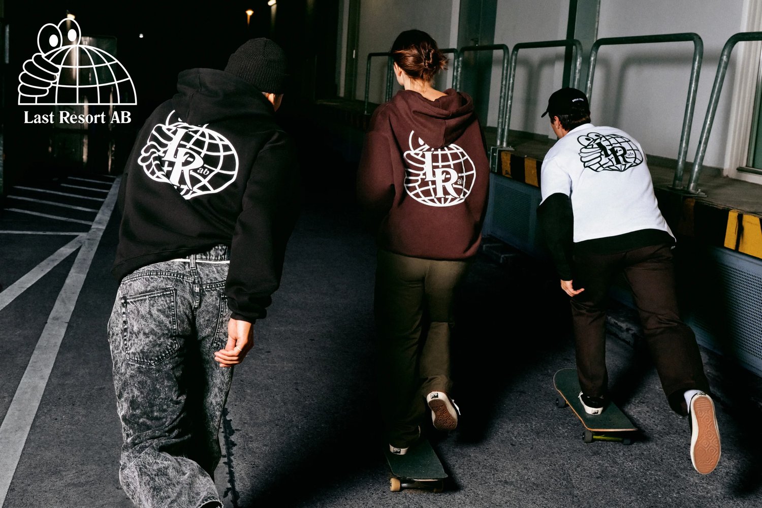 Tre individui in abbigliamento casual pattinano in un vicolo poco illuminato, mostrando felpe con cappuccio e una maglietta con un logo grafico.