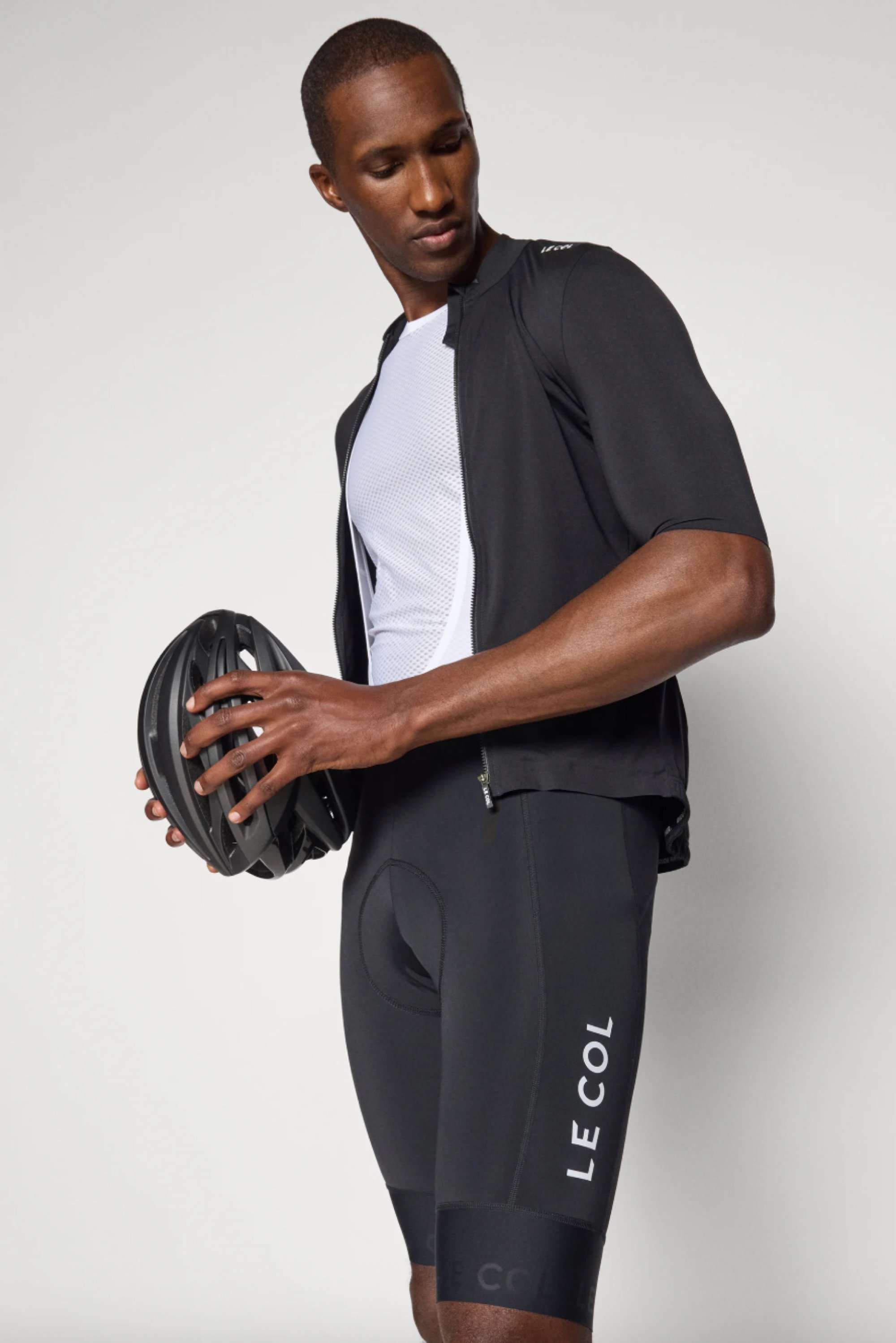 Ein Mann in sportlicher Radfahrerbekleidung hält einen schwarzen Helm in der Hand und schaut nach unten, vor einem neutralen Hintergrund. Das Outfit weist ein markenbezogenes Design auf.