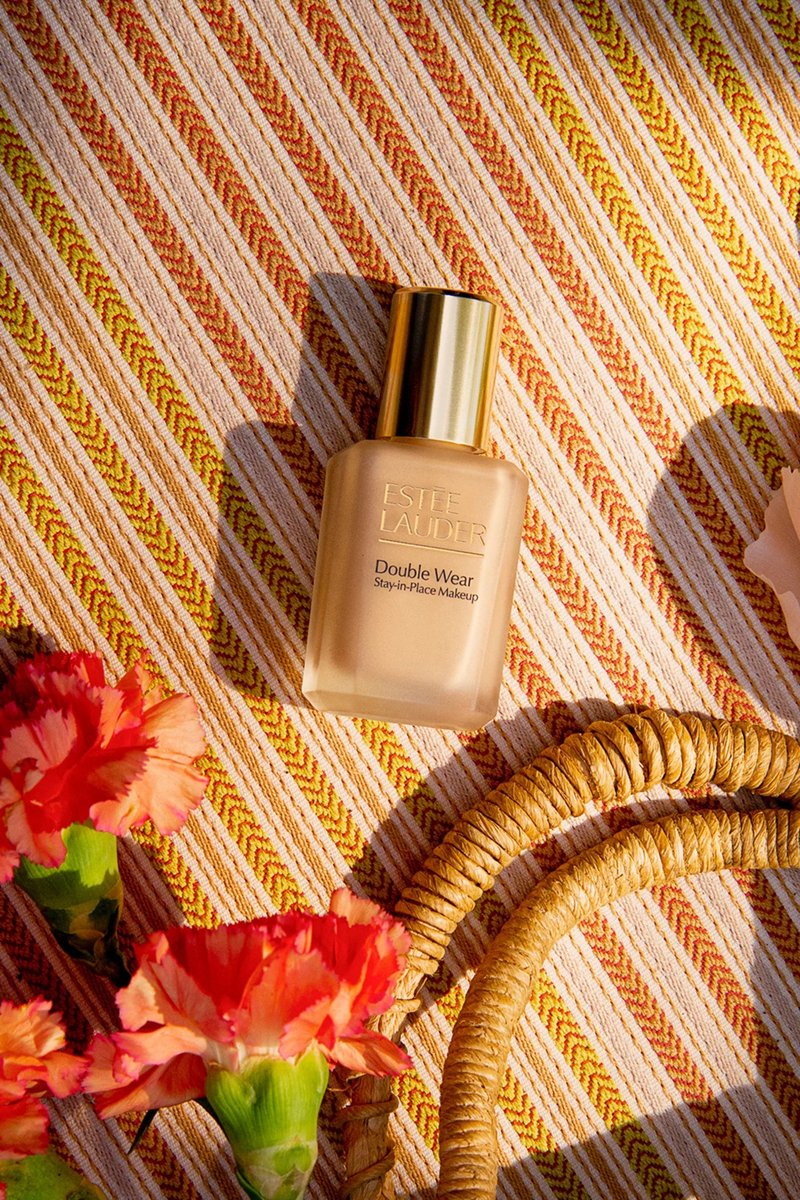 Bouteille de fond de teint Estee Lauder Double Wear sur un tissu rayé avec des œillets rouges et une anse en osier enroulée.