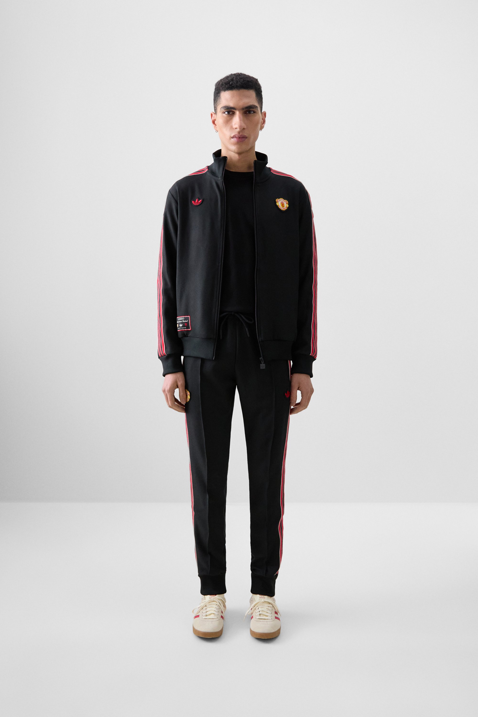 Young man standing confidently in a black tracksuit with red stripes and logo, wearing beige shoes, against a minimalistic gray background.