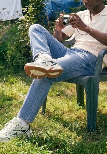 Hombre sentado al aire libre en una silla de plástico con las piernas cruzadas, sosteniendo y examinando una cámara digital compacta en un entorno de jardín.