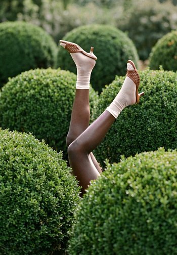 Donkerharige benen omhoog tussen groene struiken, met beige sokken en bruine open schoenen met hak in een buitentuin.