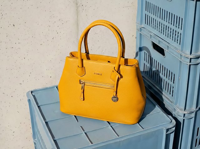 Sac à main en cuir jaune vif avec doubles poignées et poche zippée devant, posé sur des caisses en plastique bleues empilées.