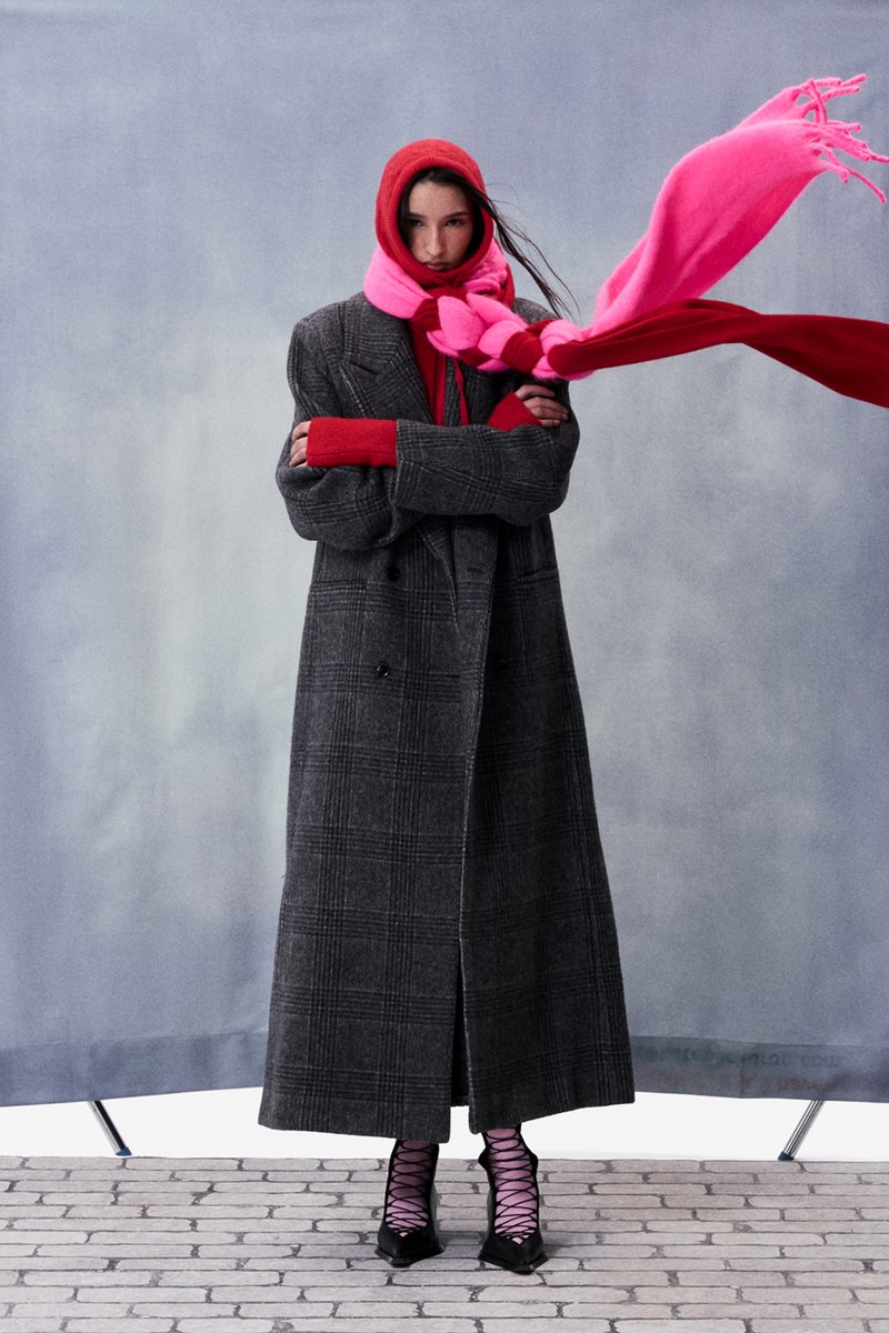 Femme portant un long manteau à carreaux gris, un sweat à capuche rouge, une écharpe rose vif et rouge flottant au vent, et des chaussures à talons hauts noires à lacets.