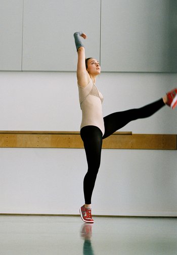 A dancer wearing a bodysuit and leggings performs a dynamic pose, one leg raised and arms extended, in a bright studio space.