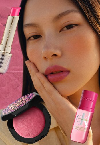 Kobieta z różową szminką i różem dotyka twarzy, otoczona różową szminką, kompaktem z różem i perfumami Calvin Klein Smooth Berry.