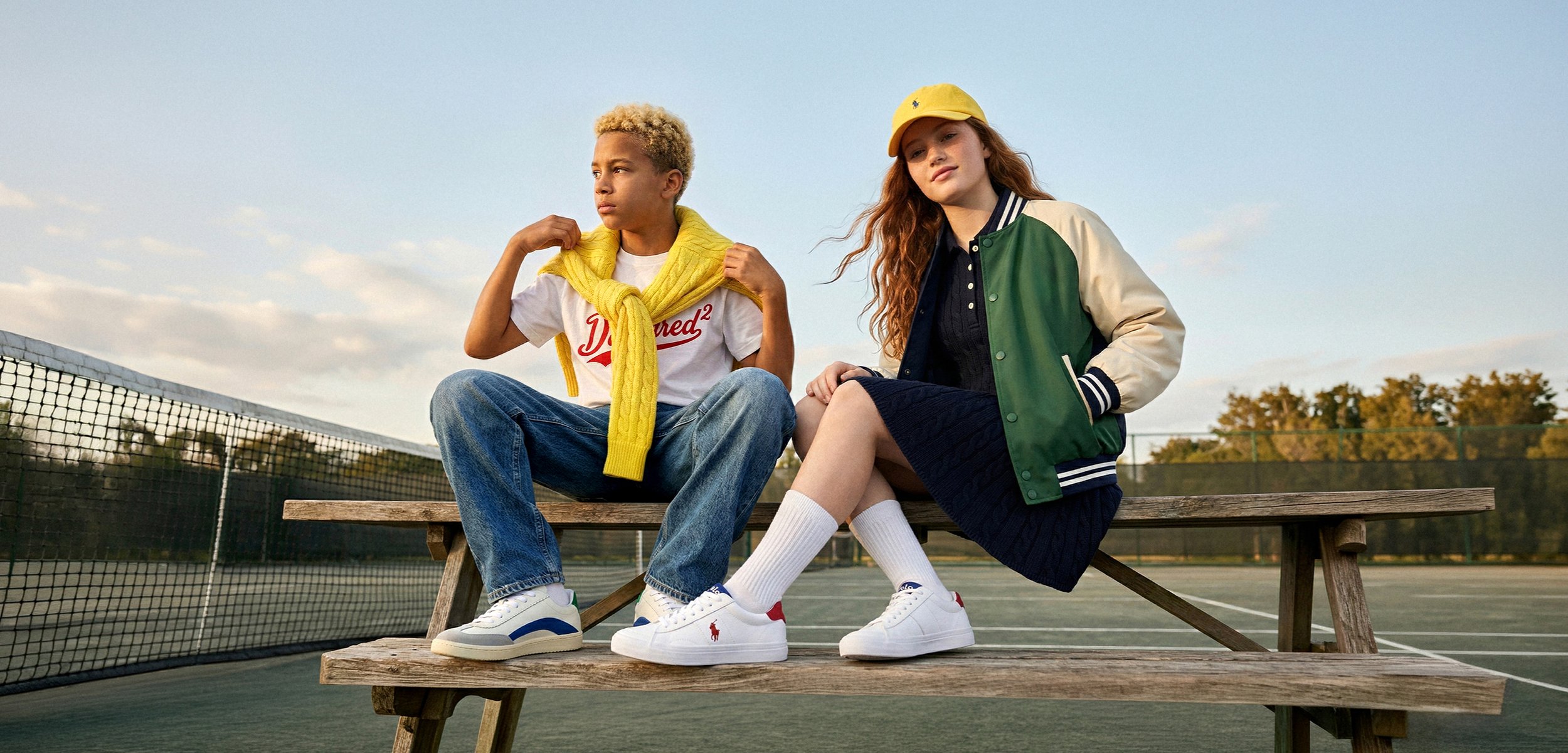 Zwei Jugendliche sitzen auf den Bänken des Tennisplatzes; einer trägt Jeans und einen gelben Pullover, der andere eine grüne Jacke und ein schwarzes Kleid.