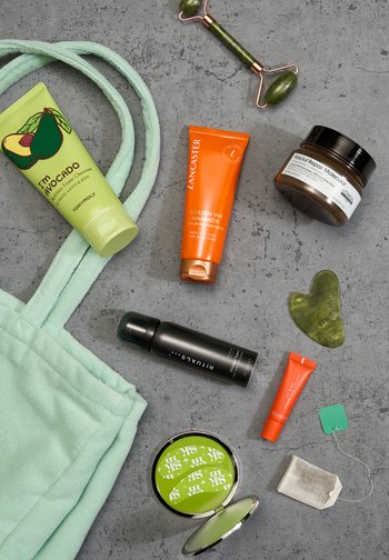 Green tote bag with skincare products, including a cleanser, tan maximizer, and a facial roller, arranged on a textured gray surface.