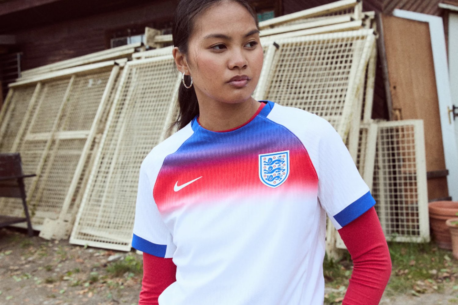 Una joven con una camiseta blanca con acentos rojos y azules, que presenta la bandera de Inglaterra, se encuentra frente a un fondo de estructuras de madera.