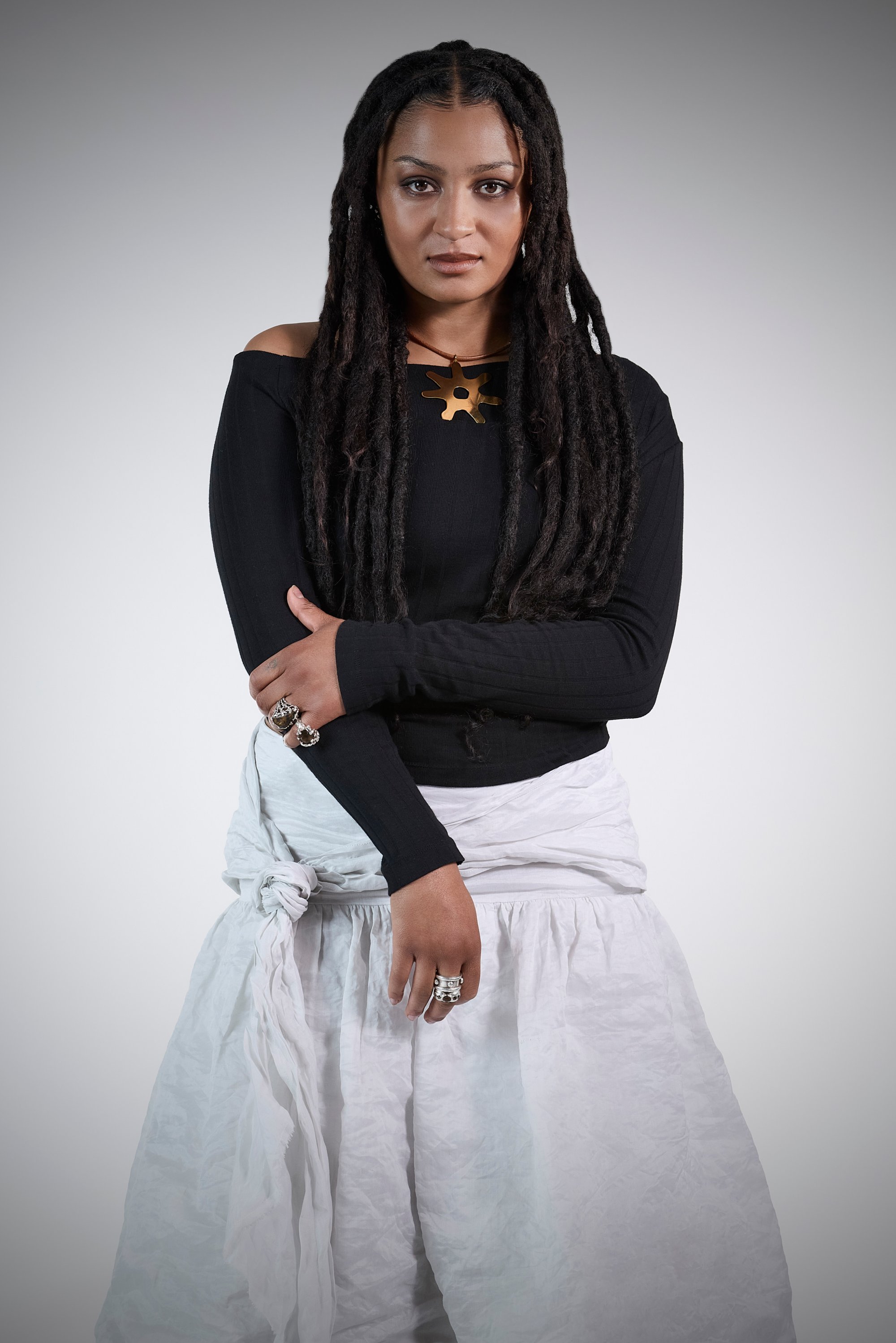 A person with long, dark dreadlocks stands confidently, wearing a black long-sleeve top, a white skirt, and layered rings.