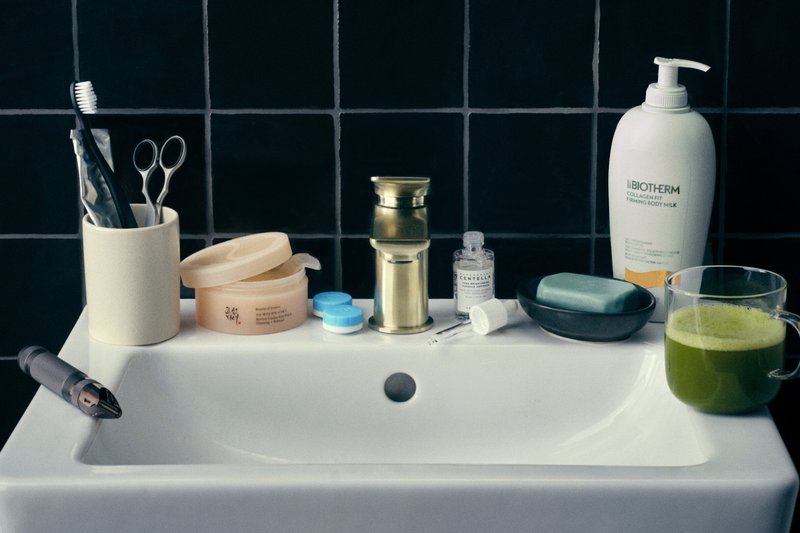 Weißes Waschbecken mit goldfarbenem Wasserhahn, Zahnbürste, Schere, Hautpflegeprodukten, Seifenstück und einem Glas grünen Saft gegen eine schwarz geflieste Wand.