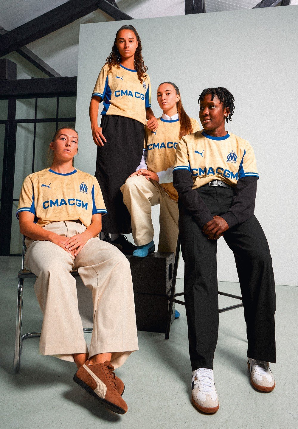 Čtyři osoby mají na sobě béžovo-modré dresy Puma s logem "CMA CGM". Jsou oblečeny do různých kalhot a obuvi v minimalistickém prostředí.