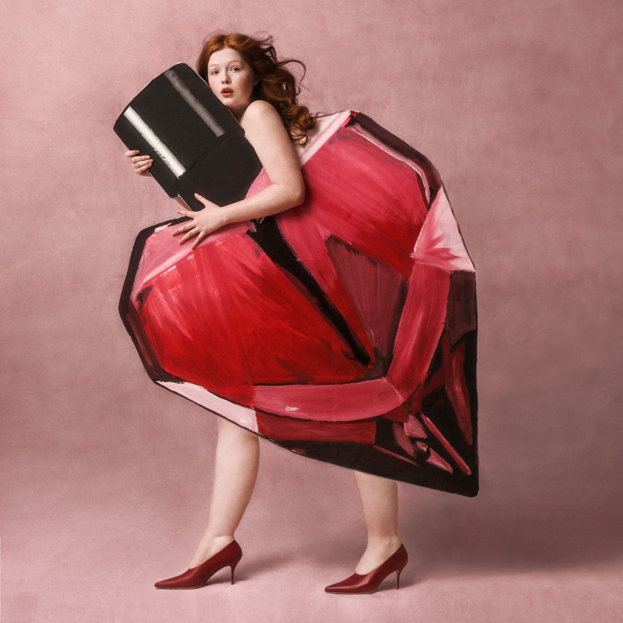 Una mujer con cabello rojo está de pie en tacones rojos, sosteniendo un gran frasco de esmalte de uñas, su vestido se asemeja a un gigantesco lápiz labial rojo. El fondo es rosa.