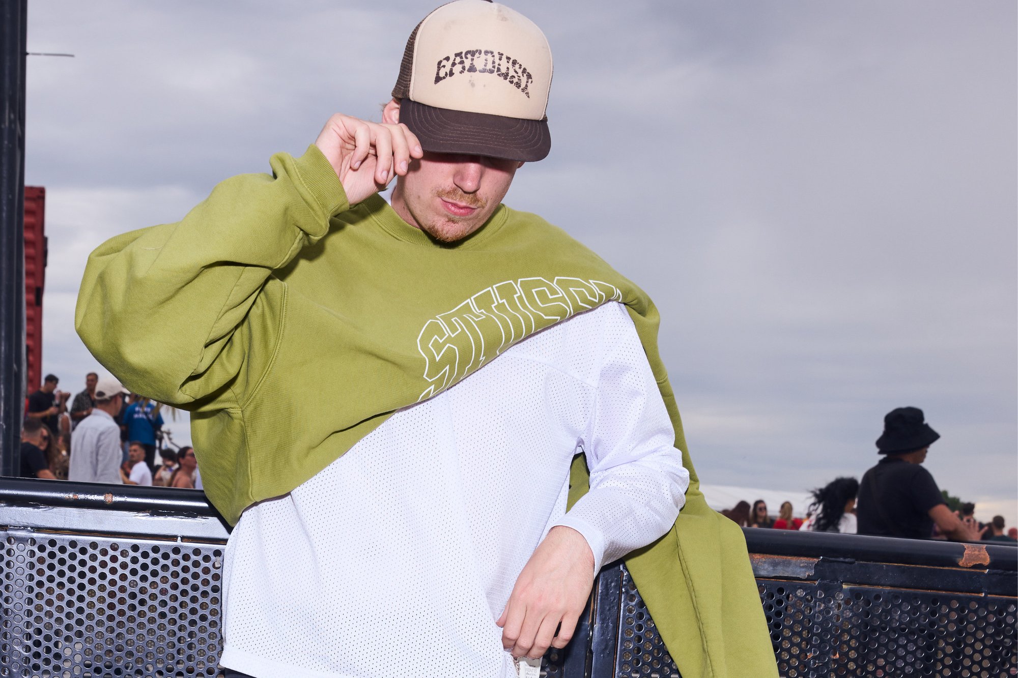 Sweat-shirt vert surdimensionné avec une insertion en maille blanche, orné d'une inscription audacieuse ; porté avec une casquette de baseball beige. L'arrière-plan comprend une foule et des éléments industriels.