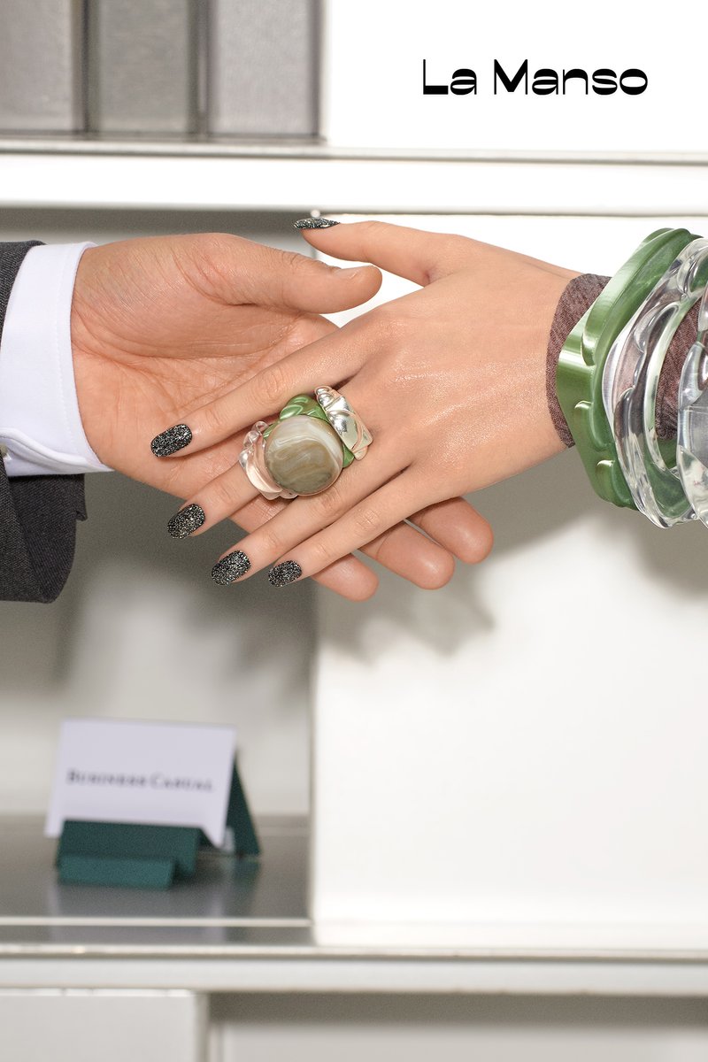 Una persona bien vestida con un traje estrecha la mano con otra persona que lleva una pulsera colorida y elegante, con objetos de oficina visibles al fondo.