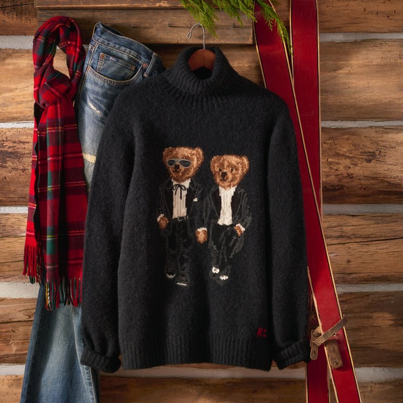 Maglione nero a collo alto con due orsacchiotti in smoking, abbinato a jeans blu, sciarpa rossa a quadri e sci vintage rossi appoggiati a una parete di legno.