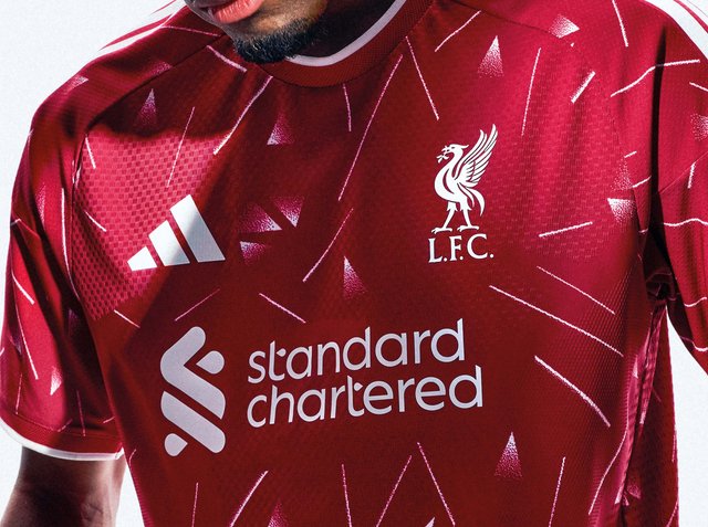 Mann trägt rotes Liverpool-Fußballtrikot mit weißen Logos, darunter Adidas, das Wappen des Liverpool FC und den Sponsor Standard Chartered.