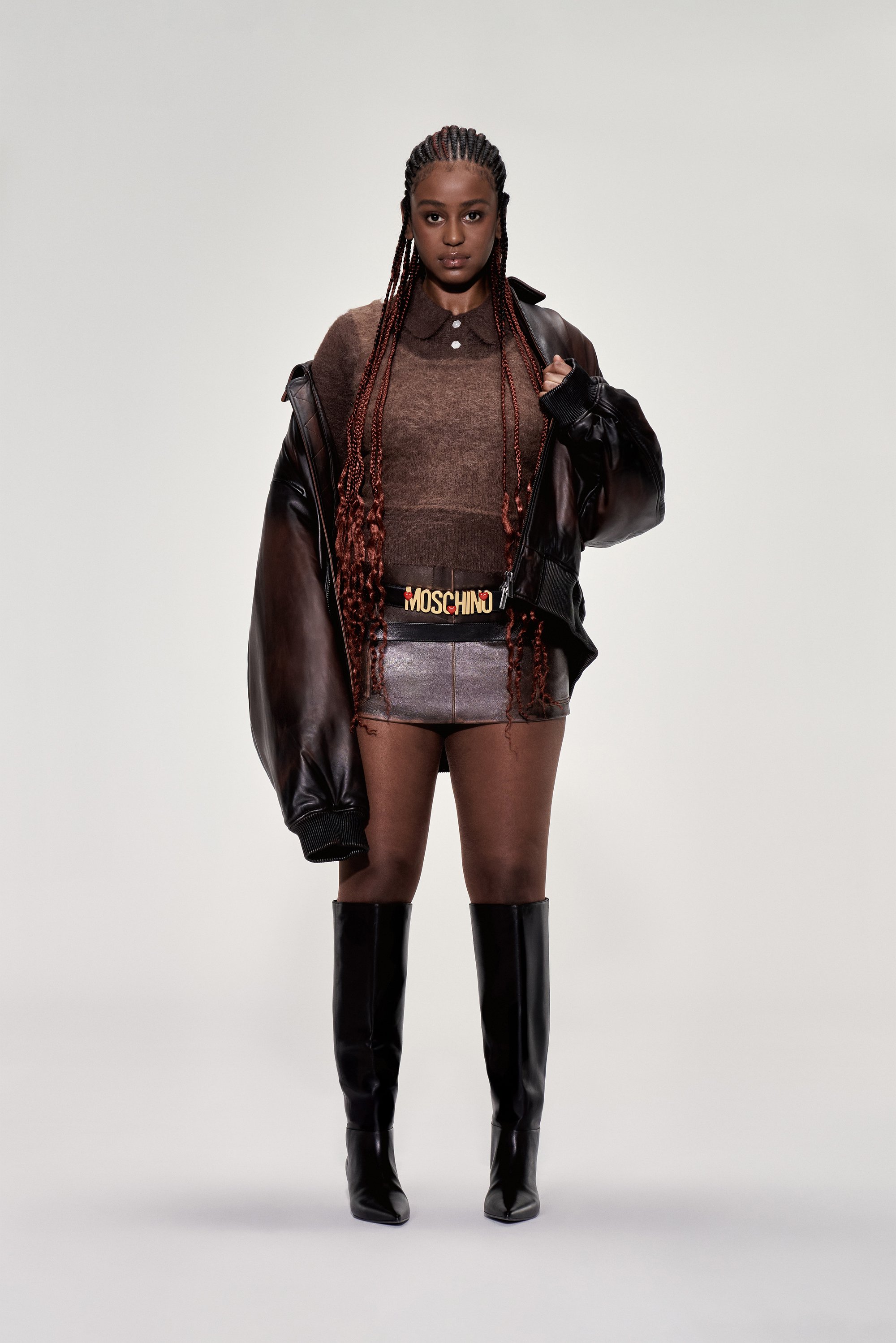 La modelo posando con confianza en un suéter marrón, chaqueta de cuero y falda corta, luciendo botas negras hasta la rodilla y un cinturón Moschino.