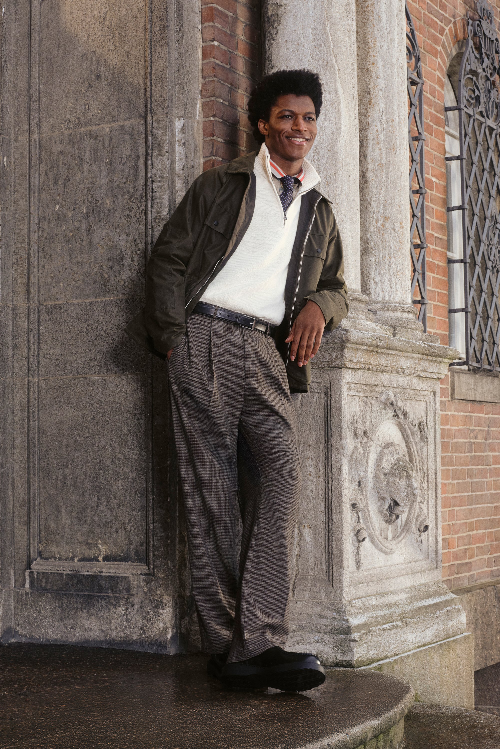A man with an afro leans against a stone wall, wearing a green jacket, white sweater, and patterned trousers, smiling confidently.