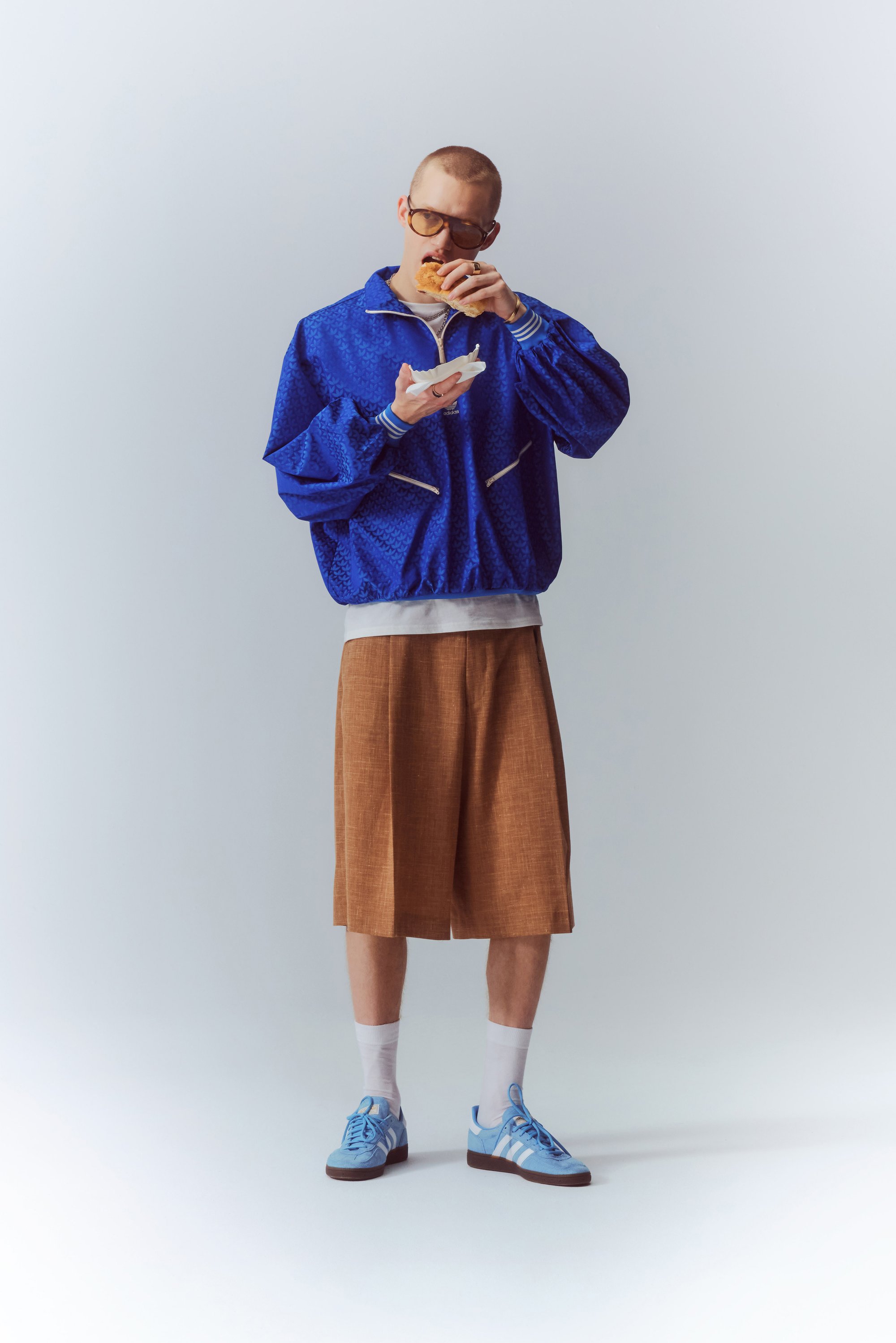 Una persona con una giacca blu, pantaloni corti marroni e sneaker blu tiene un panino, guardando concentrata mentre mastica parzialmente.