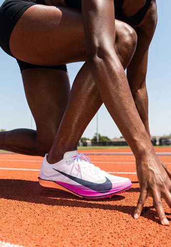 Runner in black shorts wearing white and pink Nike shoes in starting position on an outdoor track under clear sky.