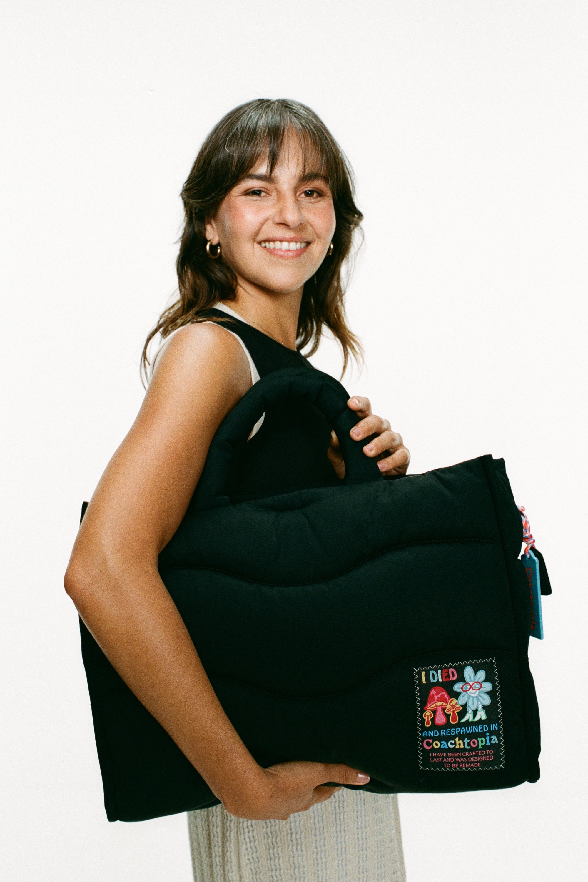 Smiling young woman holds a black padded bag with playful embroidered patches, wearing a sleeveless black top and standing against a white background.