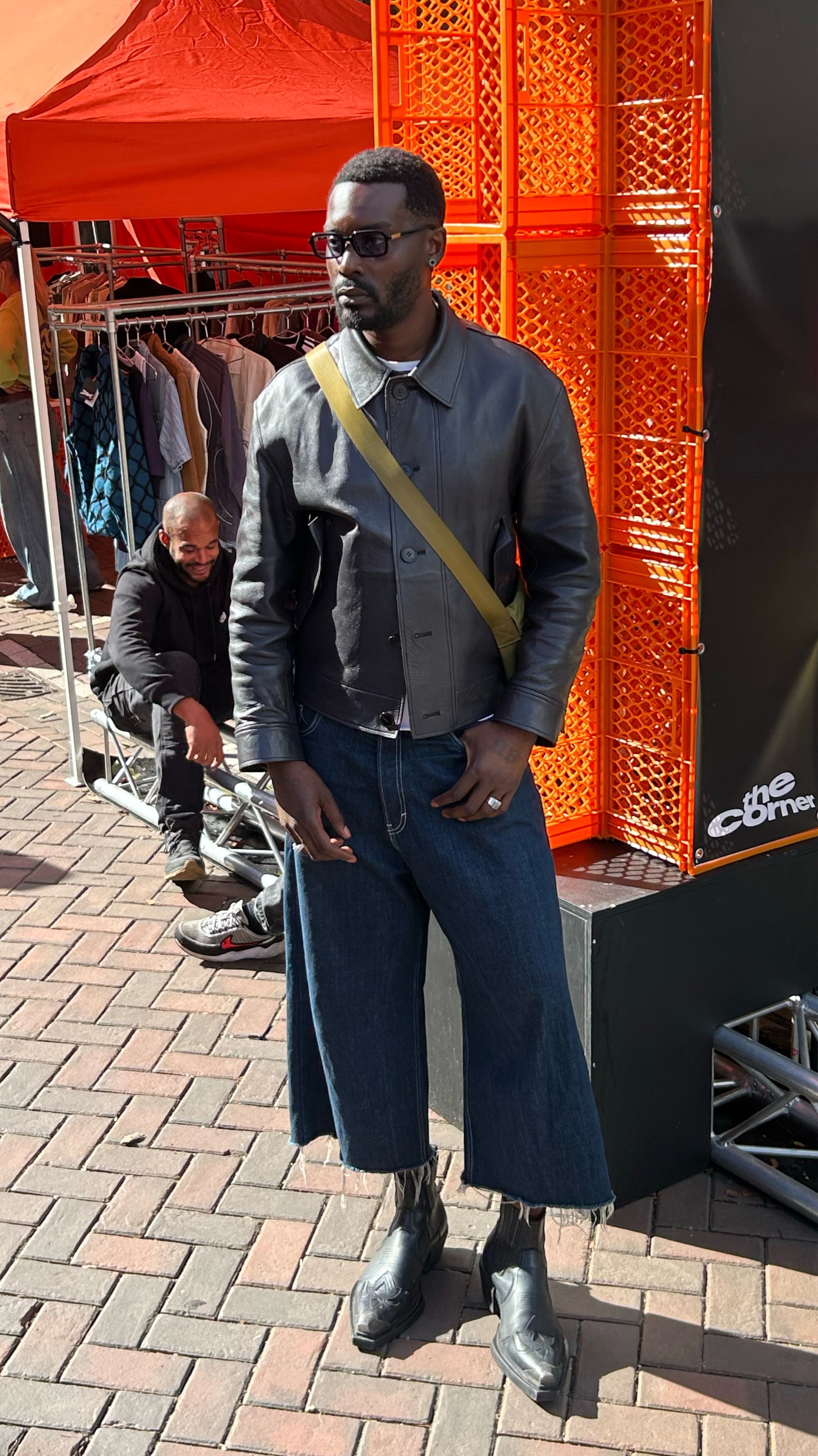 Man in een zwart leren jack, wijde cropped jeans, zwarte laarzen en een zonnebril staat op een stenen straat bij oranje displays en een kledingrek.
